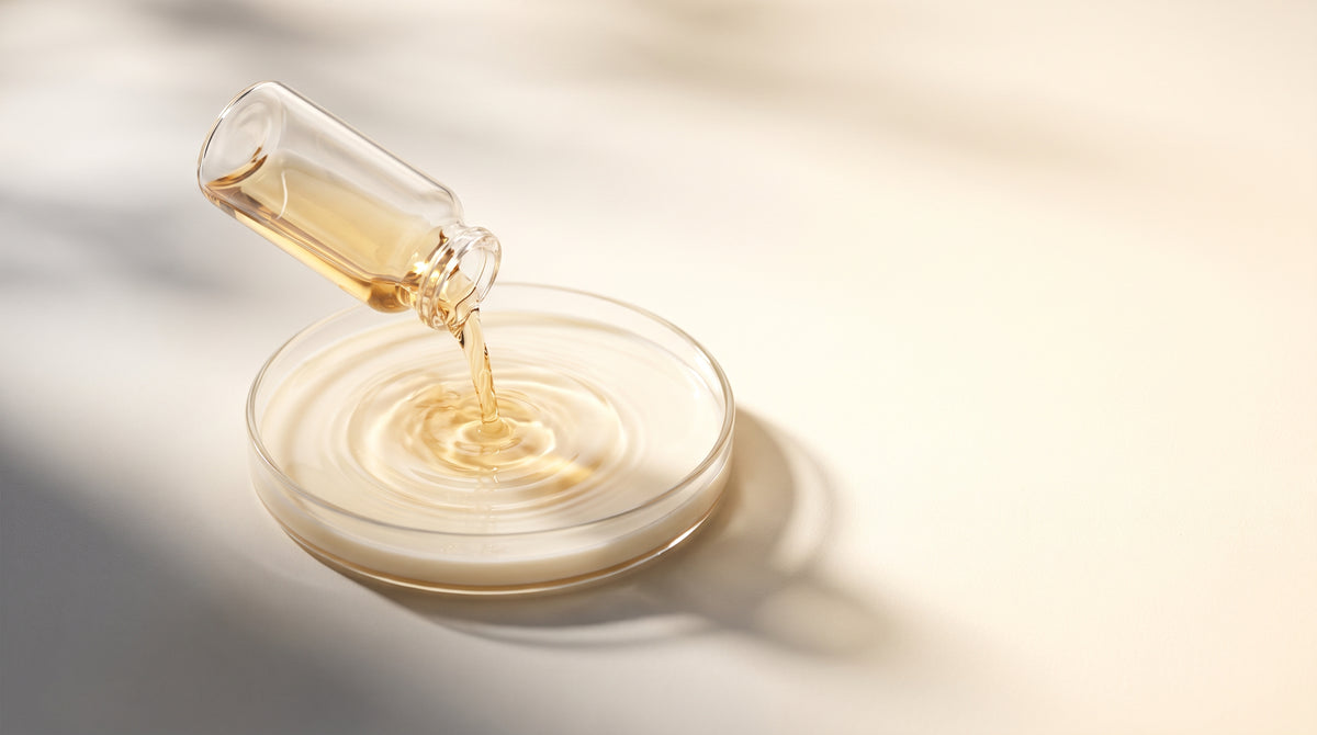 Abstract still-life of a glass vial pouring liquid into a shallow dish, warm gold and champagne tones, soft studio light