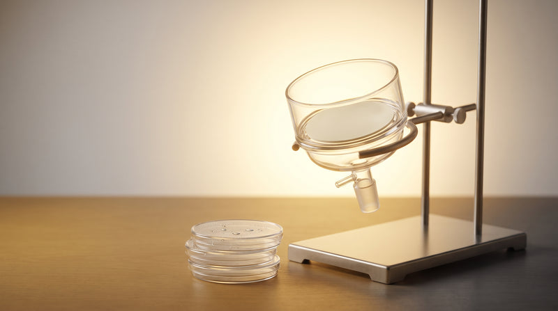 Abstract laboratory still life of a glass membrane filtration apparatus and petri dishes in warm gold and ivory tones