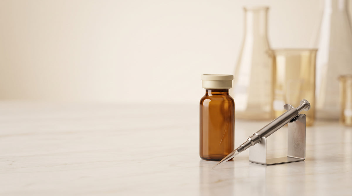 Abstract still-life of an amber laboratory vial and micropipette on a pale marble bench, warm gold and ivory tones
