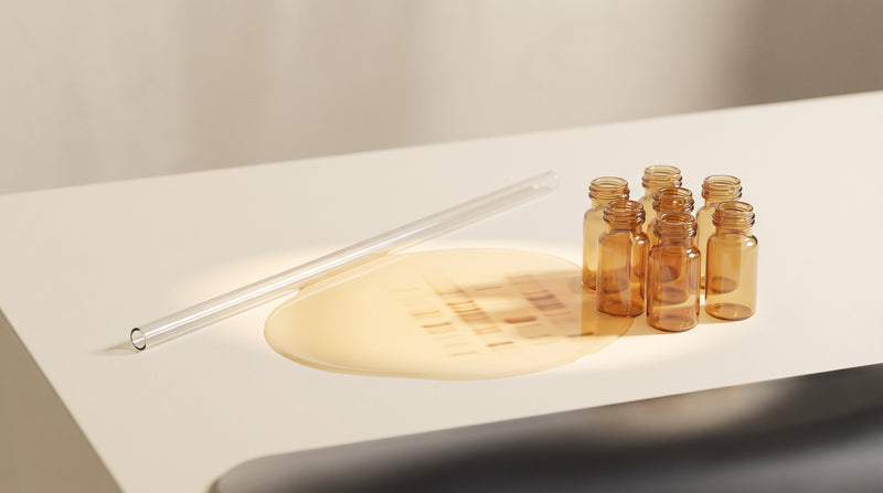 Abstract still life of a slender glass capillary tube and amber laboratory vials in warm gold and champagne tones with soft studio lighting