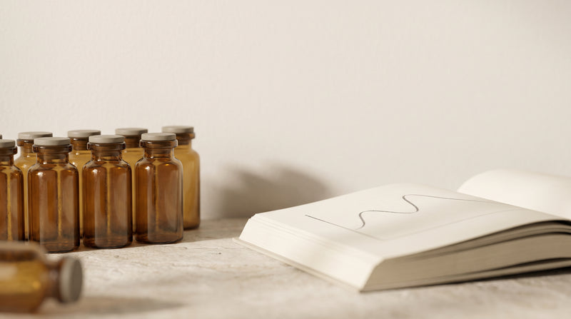 Amber laboratory glass vials beside an open reference book with a faint chromatography curve, warm gold and ivory studio lighting