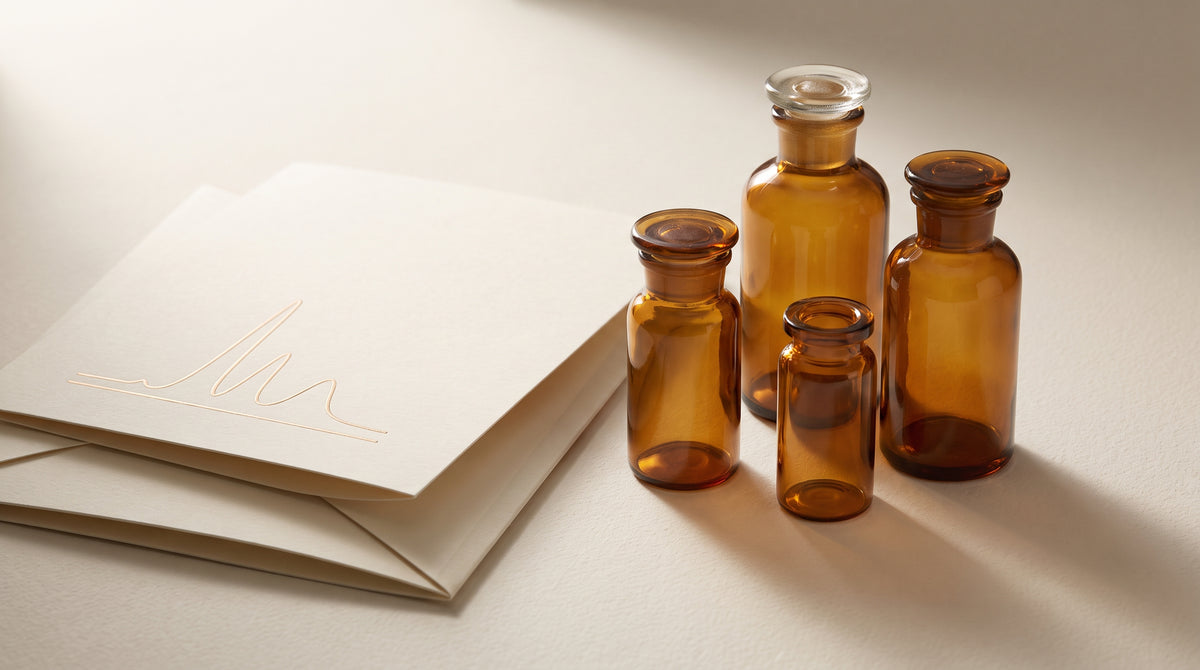 Amber laboratory glass vials of varying sizes beside an embossed ivory document with a subtle chromatography-curve motif, softly lit in warm gold and champagne tones